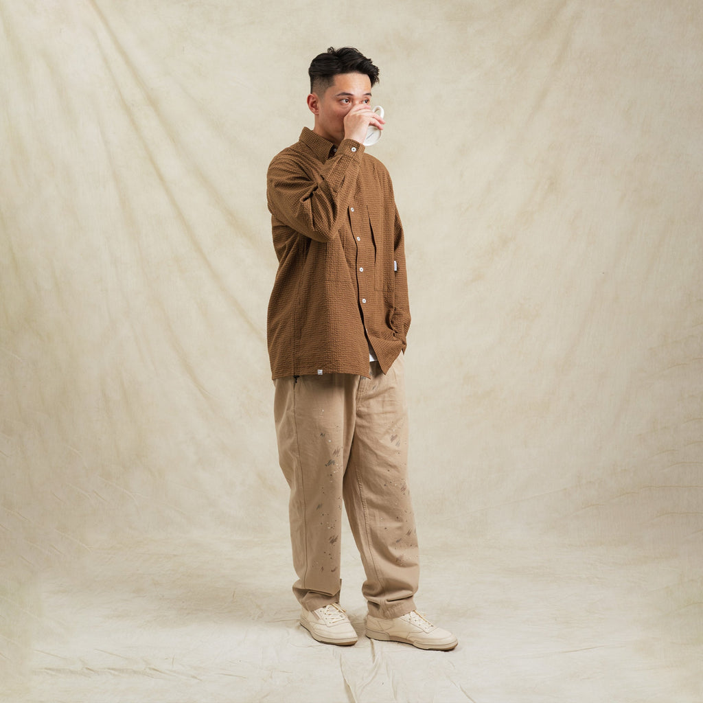 Man wearing a brown jacket and beige pants holding a mug against a beige background