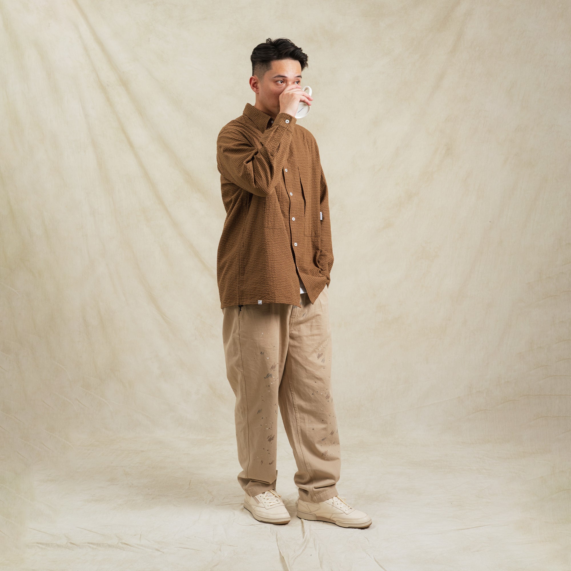 Man wearing a brown jacket and beige pants holding a mug against a beige background